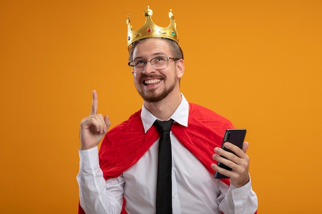 Бесплатное фото Впечатленный молодой супергерой в галстуке и короне в очках держит телефон и указывает вверх на оранжевом фоне