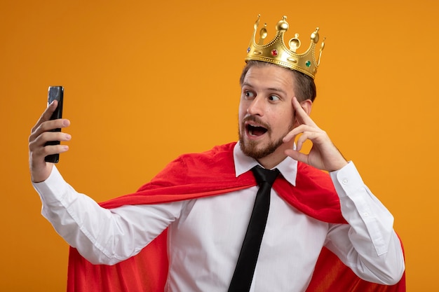 Impressed young superhero guy wearing tie and crown take a selfie isolated on orange