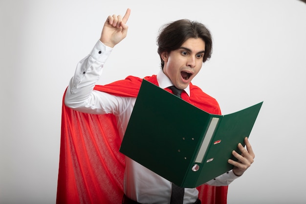 Free photo impressed young superhero guy wearing tie holding and looking at folder points at up isolated on white background