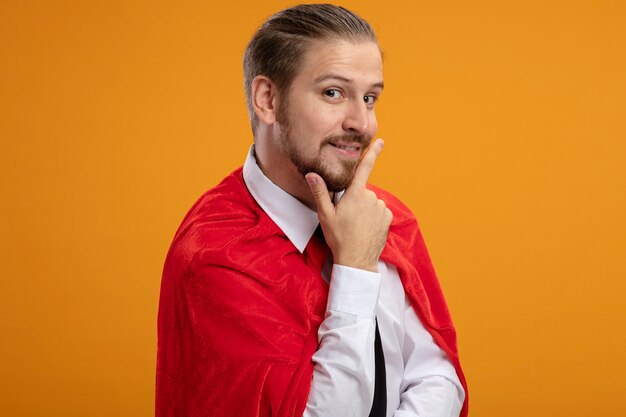 Impressed young superhero guy wearing tie putting hand under chin isolated on orange