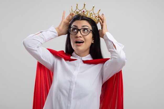 Impressed young superwoman wearing glasses and crown touching crown looking up isolated on white wall