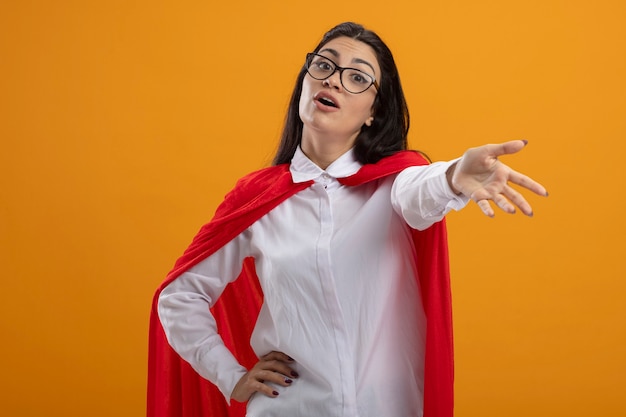 Impressed young superwoman wearing glasses looking at front stretching out hand towards front keeping another one on waist isolated on orange wall