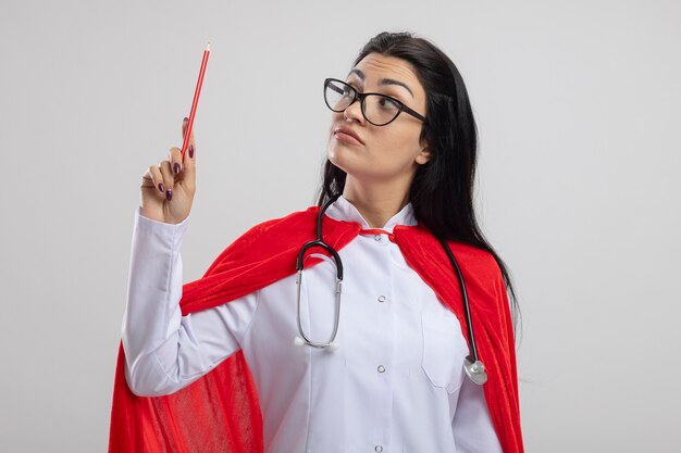 Impressed young superwoman wearing glasses and stethoscope holding and looking at red pencil isolated on white wall