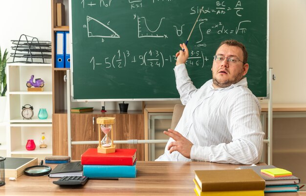 impressed young teacher wearing glasses sitting at desk with school supplies in classroom looking at front pointing at chalkboard with pointer stick