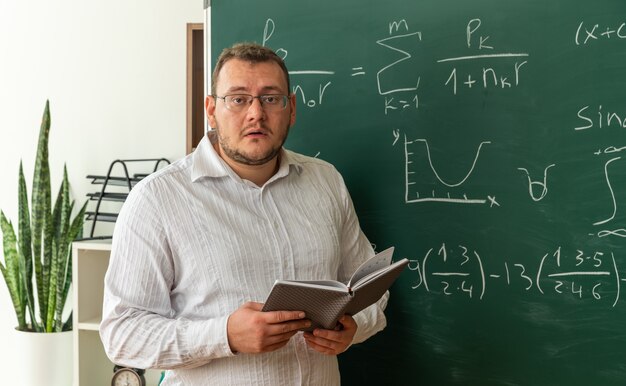 impressed young teacher wearing glasses standing in front of chalkboard in classroom holding open notepad looking at front