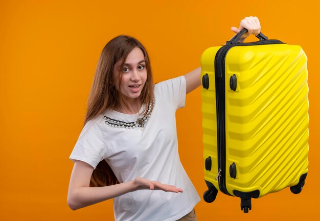 Impressed young traveler girl raising suitcase and pointing with hand at it on isolated orange space