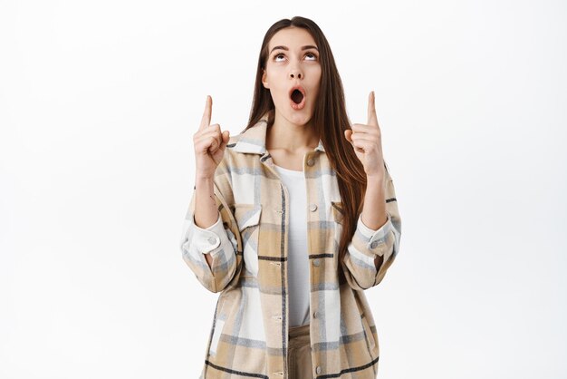 Impressed young woman drop jaw say wow and stare at promotional text with amazed face expression pointing fingers up at cool shop offer standing over white background