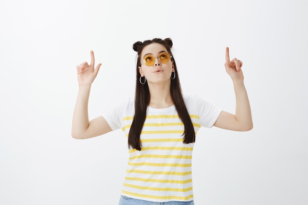 Впечатленная молодая женщина позирует в солнцезащитных очках на белой стене