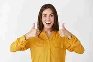 Free photo impressed young woman showing thumbs up and smiling amazed, praising something cool, standing over white wall