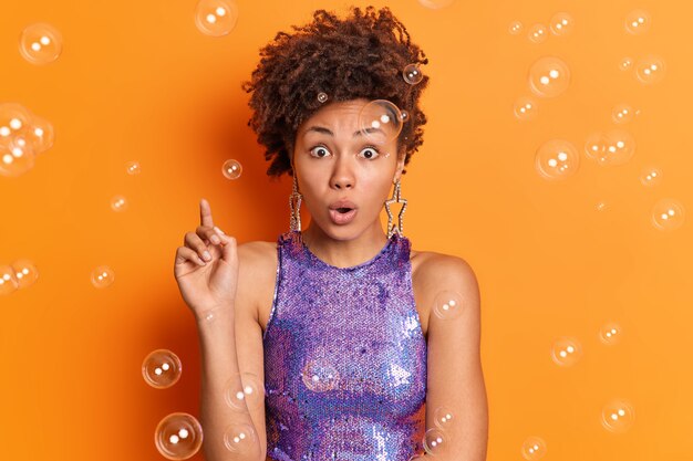 Impressed young woman with curly hair looks stunned at camera cannot believe her eyes wears stylish clothes isolated over orange wall soap bubbles flying around