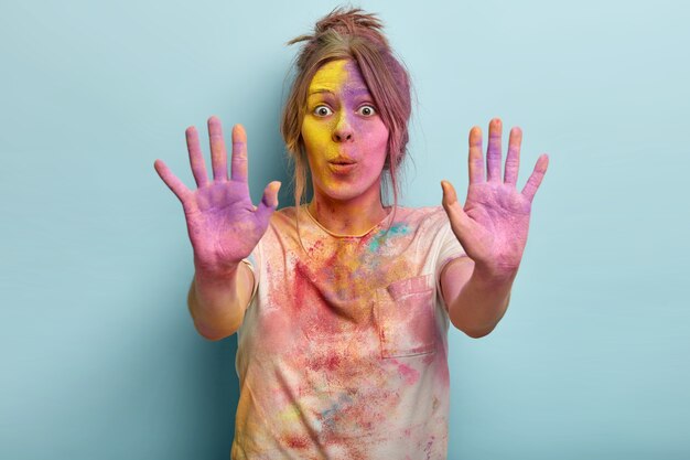 Impressive European female has surprised facial expression, shows both colored palms, smeared face with powder, stares from shock has dirty t shirt plays with colors on Holi festival. Reaction concept