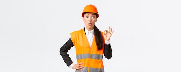 Imressed and surprised asian female construction manager pleased with amazing quality standing in safety helmet and reflective jacket showing okay gesture and say wow white background