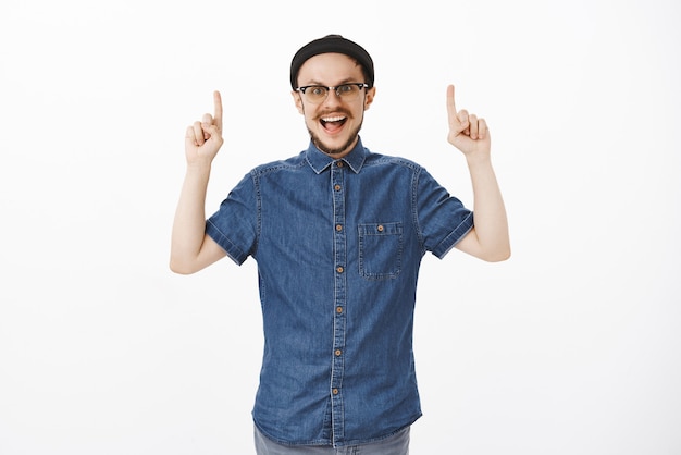 Imrpessed and amazed joyful european male with beard in glasses and trendy beanie raising hands pointing up and smiling broadly from excitement and thrill happy like child during tourist travel