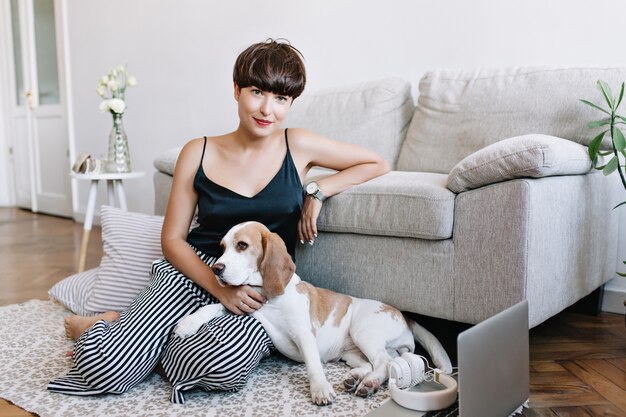 Incredible brunette lady wears elegant wristwatch posing beside sofa stroking cute beagle