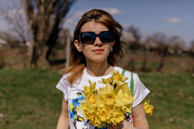 Incredible pretty girl in sunglasses dressed white tshirt is holding bouquet of flowers outside in sunny warm day on background of blue sky