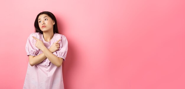 Indecisive asian woman cant decide pointing fingers sideways and look pensive aside making choice st