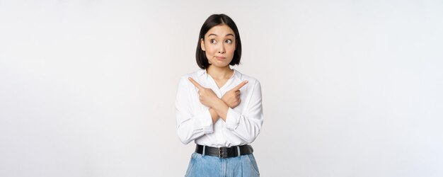 Нерешительная азиатка, указывающая пальцами в сторону, указывающая пальцами и выглядящая невежественной, сбитой с толку выбором, стоящим на белом фоне