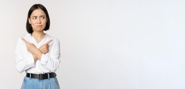 Indecisive asian woman pointing fingers sideways pointing fingers and looking clueless confused with choices standing over white background