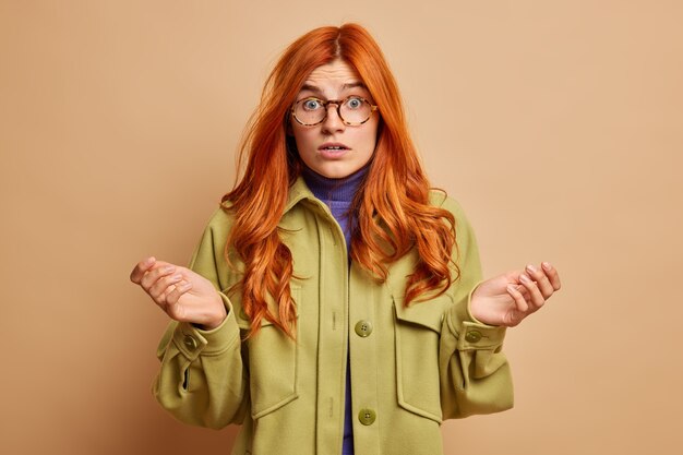 Indecisive confused redhead woman spreads palms sideways and feels confused stares with shocked expression wears fashionable outerwear.