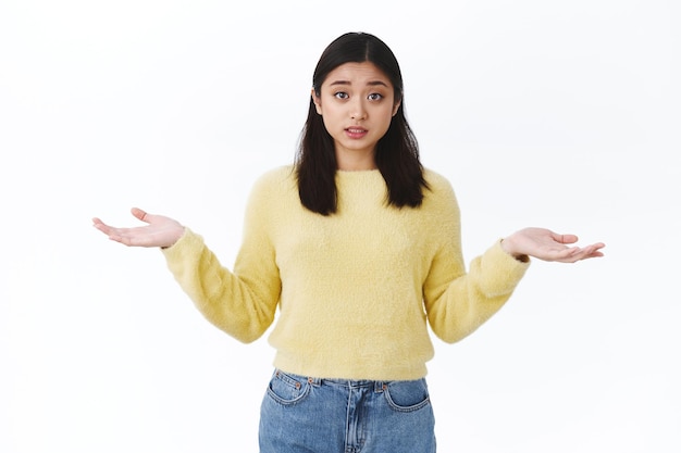 Indecisive cute asian girl asking your advice as dont know what do, shrugging spread hands sideways and looking camera questioned