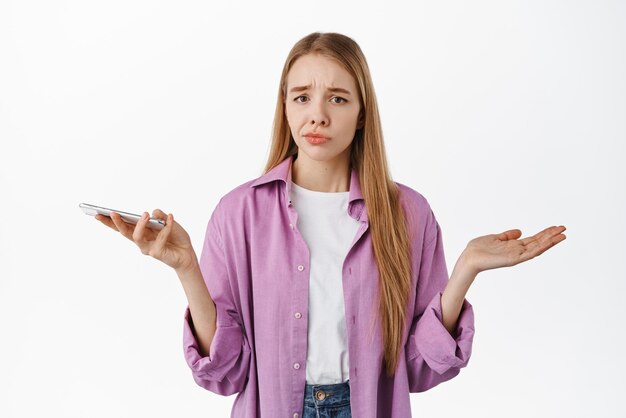 Indecisive cute girl holding smartphone spread hands sideways and shrugging confused dont know nothing what to do standing over white background