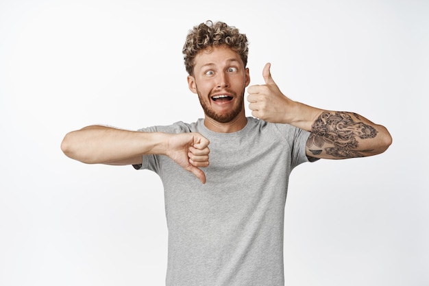 Free photo indecisive funny guy making crazy eyes showing tongue thumbs up and thumbs down cant make decision rate something give feedback like or dislike signs white background