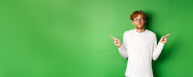 Free photo indecisive funny guy with red hair pointing fingers sideways staring confused at camera and showing