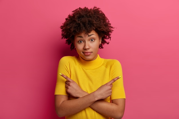 Indecisive girl picks from two choices, looks questioned and troubled, crosses hands across chest, hesitates between suggested products, wears yellow t shirt, isolated over crimson wall