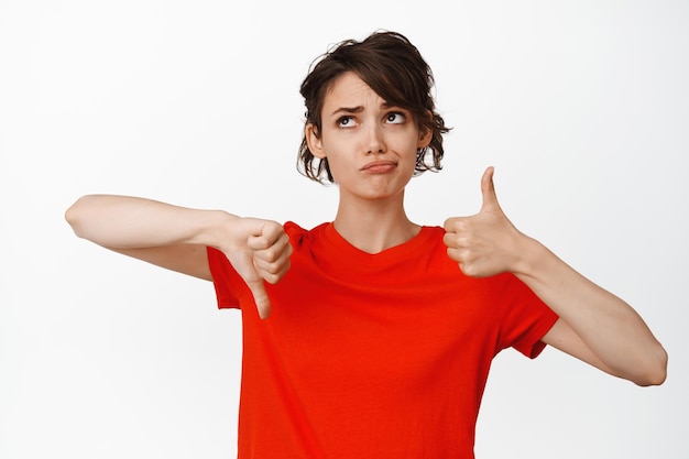 Indecisive girl showing thumbs up and thumbs down, looking puzzled, cannot make choice, weighing pros and cons, white background