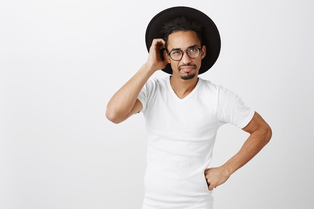 Indecisive handsome african-american man in glasses and hat scratch head puzzled, looking confused