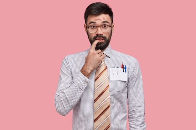 Нерешительный интеллектуал держит палец у рта, имеет густую бороду, одет в строгую рубашку и галстук, носит большие очки.