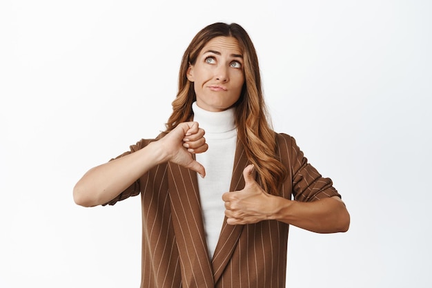 Free photo indecisive middle aged corporate woman weighing decision showing like dislike gesture thumbs up and thumbs down looking up and thinking white background
