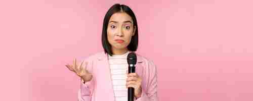 무료 사진 우유부단한 신경질적인 아시아 비즈니스 여성이 마이크를 으쓱하고 분홍색 배경에 정장을 입고 마이크를 들고 아무 생각 없이 서 있는 모습을 보고 있습니다