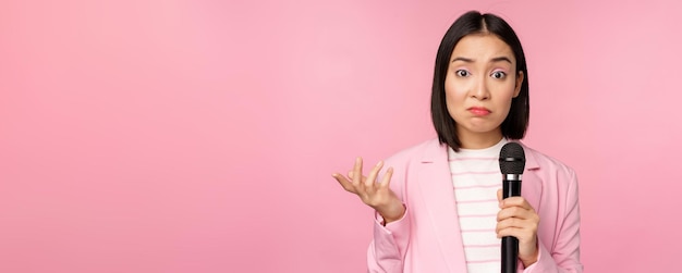 우유부단한 신경질적인 아시아 비즈니스 여성이 마이크를 으쓱하고 분홍색 배경에 정장을 입고 마이크를 들고 아무 생각 없이 서 있는 모습을 보고 있습니다
