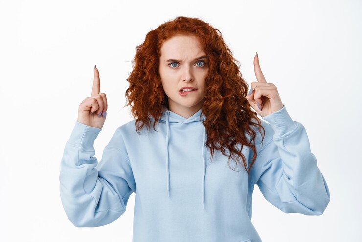 Free Photo indecisive redhead girl need help with decision pointing fingers up while biting lip and stare doubtful at camera standing in hoodie against white background