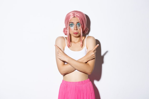 Indecisive silly female model in pink wig, with colorful makeup for halloween party, pointing sideways