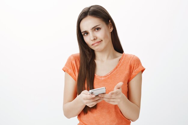 Indecisive and uncertain cute brunette girl shrugging while using smarpthone, smirk unsure