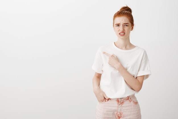 Indecisive and unsure redhead girl frowning perplexed and pointing left