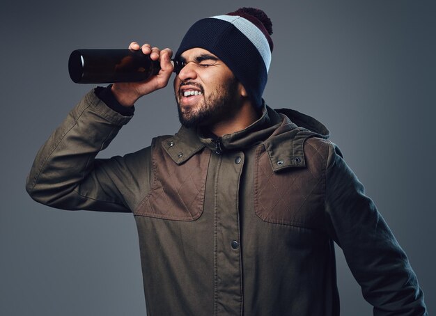 An Indian bearded male dressed in a warm jacket and a hat looking through beer bottle.