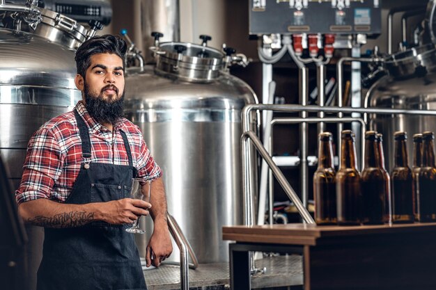 地ビール醸造所でクラフトビールを提示するインドのひげを生やした入れ墨の流行に敏感な男性メーカー。