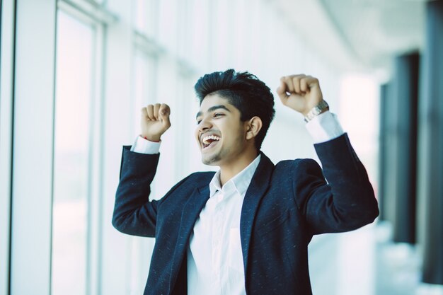 Индийский бизнесмен в костюме, выражая успех выиграть жест возле окна в офисе
