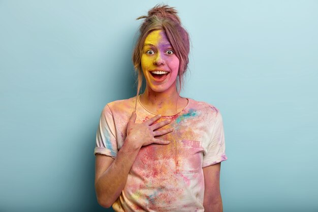 Indian festival concept. Impressed beautiful European female keeps hand on chest, looks in joy, covered with colorful dyes, has dirty smeared t shirt, has fun with colors, isolated on blue