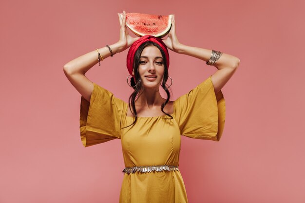 Indian girl with black wavy hair in headband and yellow fashionable clothes holding watermelon on her head on pink wall