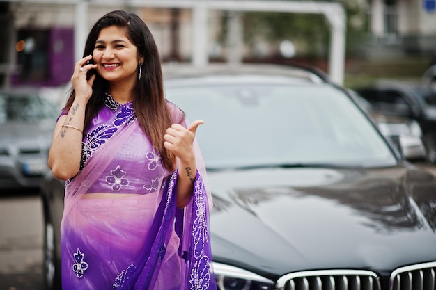 전통적인 바이올렛 사리를 입은 인도 힌두교 소녀가 거리에서 검은색 비즈니스 SUV 차량에 맞서 포즈를 취하고 휴대전화로 말하는 것은 엄지손가락을 치켜드는 모습을 보여줍니다.