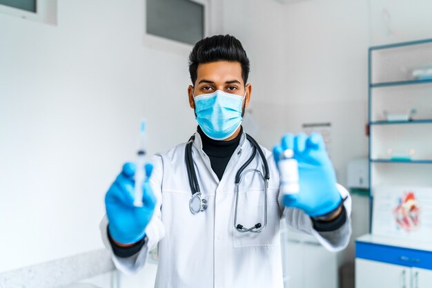 An Indian male doctor in a protective mask and blue glove holds an injectionnd vaccine in his hands and points