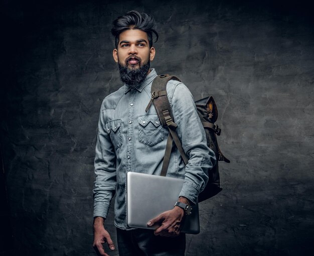 The Indian male student holds laptop and backpack over grey studio background.