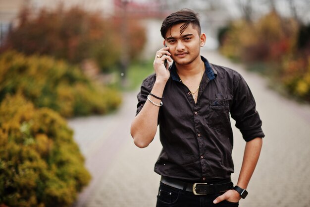 茶色のシャツを着たインド人男性が屋外でポーズをとり、携帯電話で話します