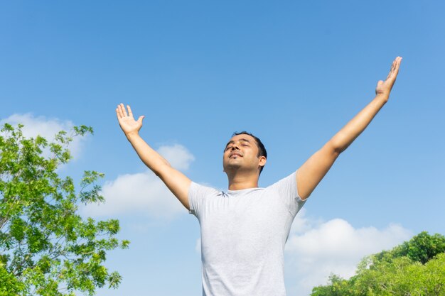 Индийский человек, концентрируясь и поднимая руки на открытом воздухе с голубым небом и зелеными ветвями деревьев