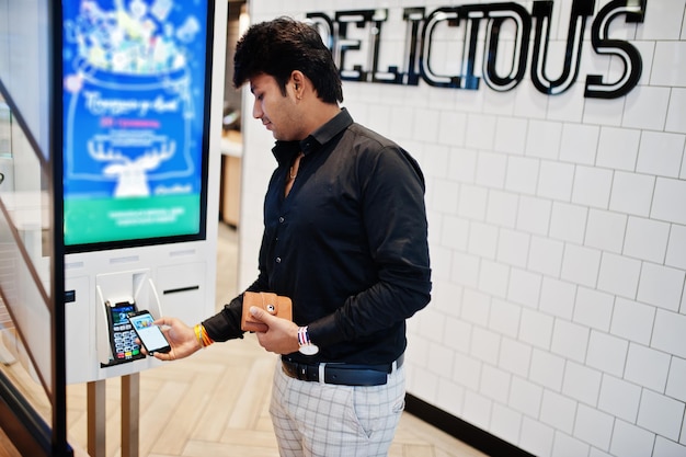 Indian man customer at store place orders and pay by contactless credit card on mobile phone through self pay floor kiosk for fast food payment terminal Pay pass