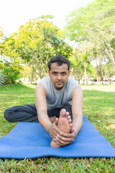 Uomo indiano che fa piegare in avanti testa a ginocchio all'aperto sulla stuoia in parco con gli alberi
